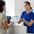 Mitarbeiterin in blauer medizinischer Kleidung reicht einer Patientin ein Formular zur Befragung