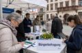 Bürgerbefragung vor einem Rathaus mit Papierfragebogen und Tablet als Beispiel für hybride Befragungen in Behörden