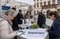 Bürgerbefragung vor einem Rathaus mit Papierfragebogen und Tablet als Beispiel für hybride Befragungen in Behörden 